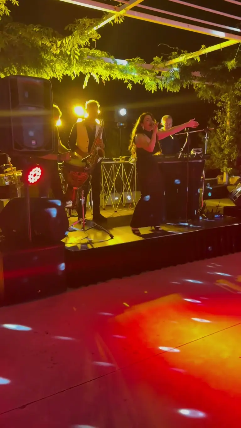 Live band che suona sul palco sotto un pergolato illuminato: voce, sax e percussioni con diffusori e monitor ai lati