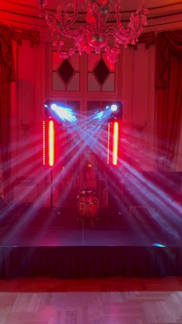 Sala da ballo con lampadario, barre LED rosse e due teste mobili che incrociano i fasci sopra il palco con la batteria