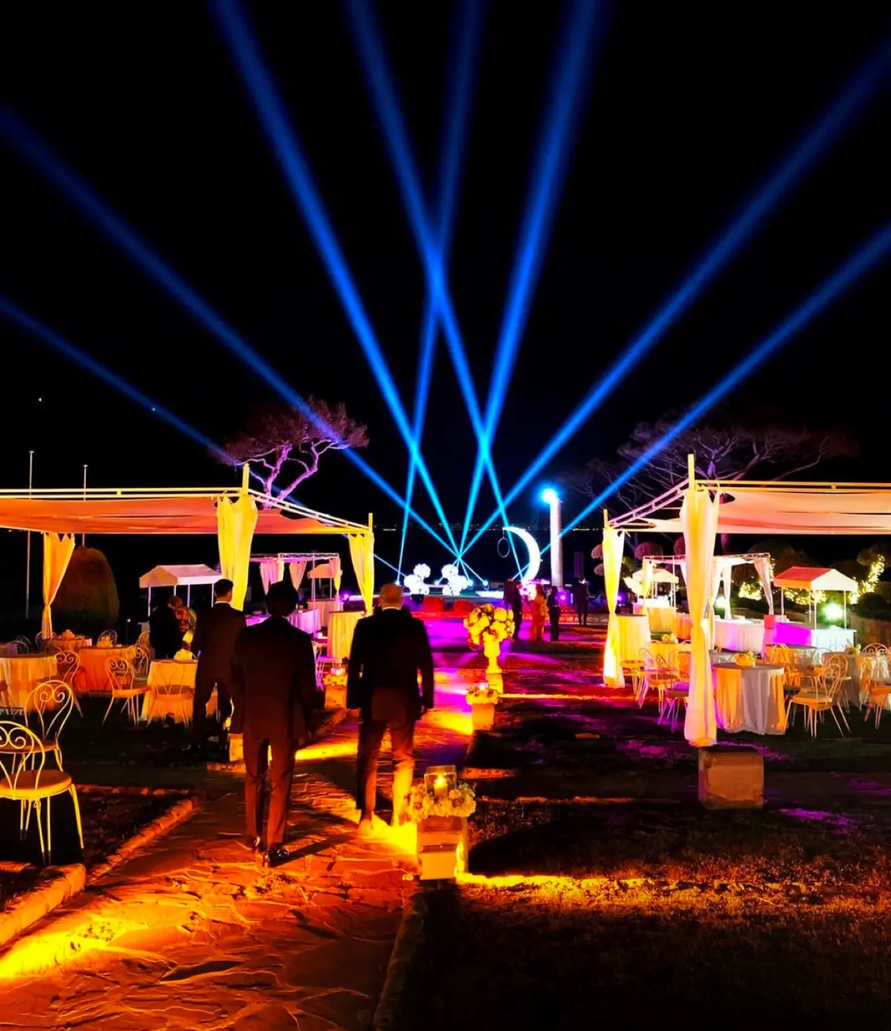 Cena di gala all'aperto con fasci blu nel cielo e illuminazione calda sui tavoli