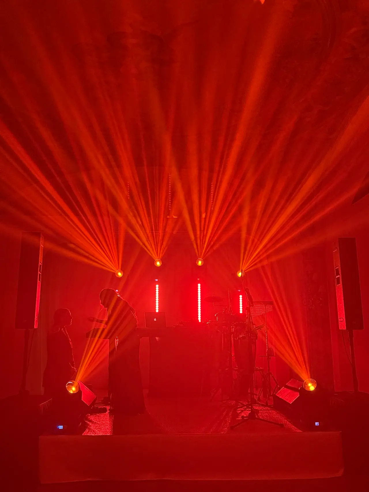 Palco di una live band con teste mobili e fasci rossi nella foschia, torri audio ai lati