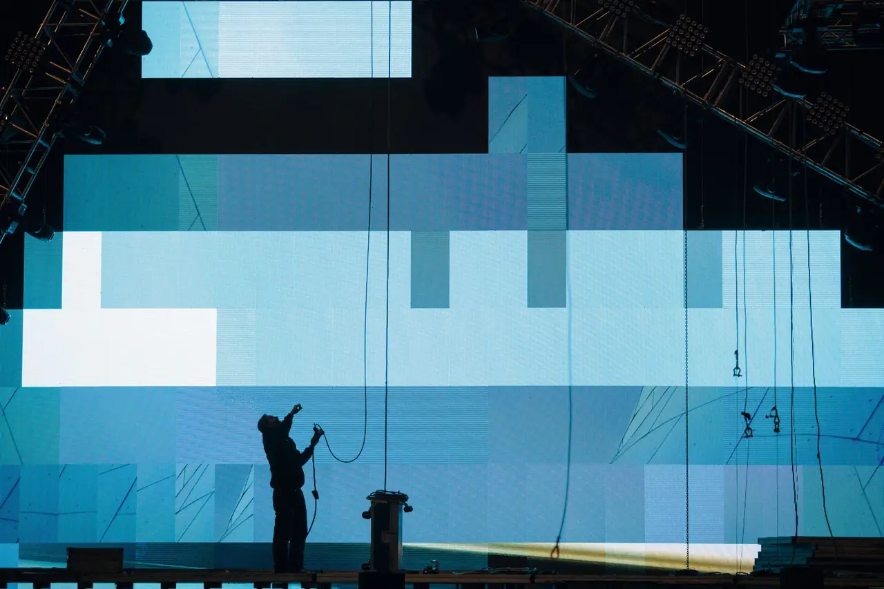 Tecnico in controluce davanti a un grande ledwall durante il montaggio, con truss e cavi ancora a vista