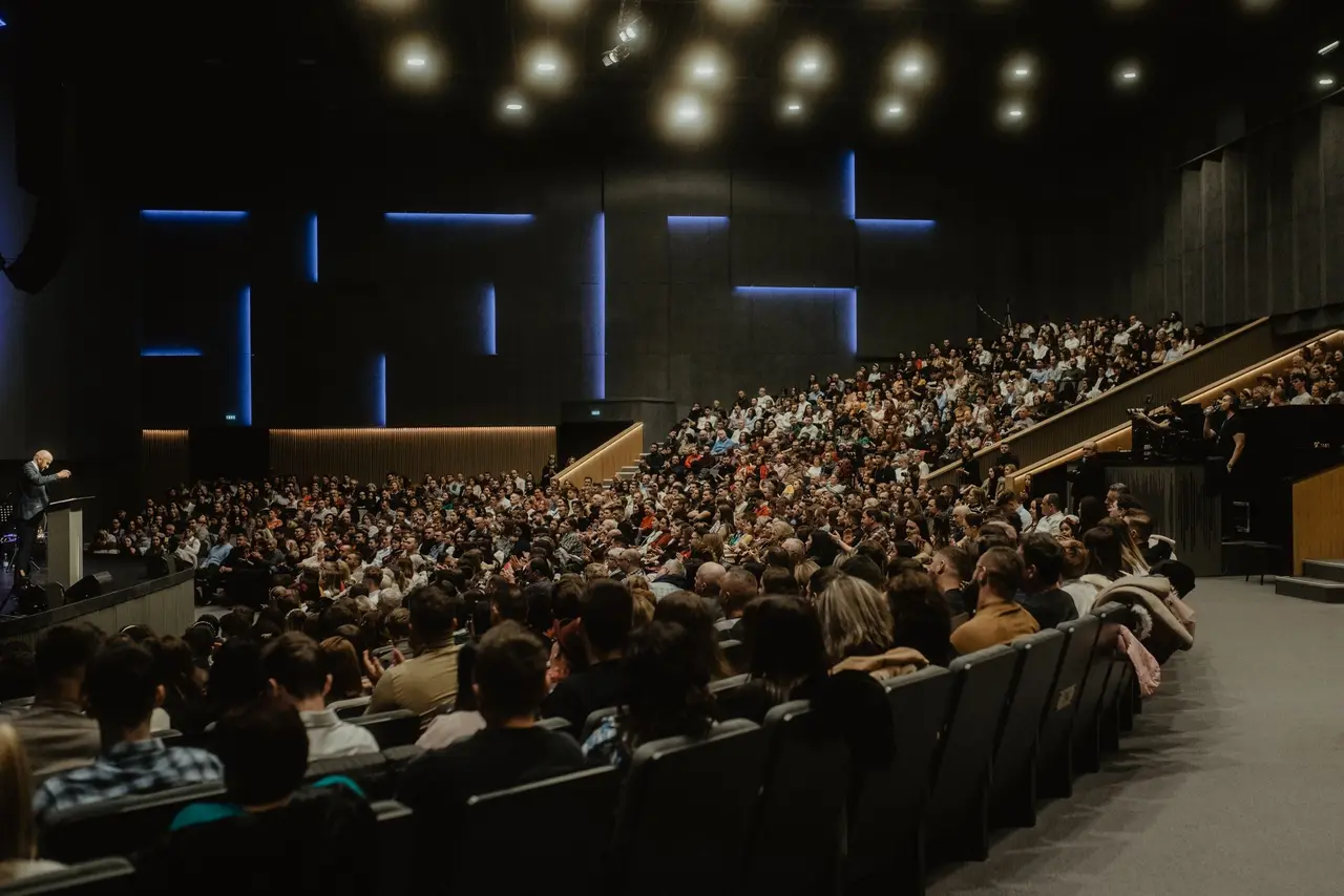 Platea gremita di un auditorium durante un convegno, pareti scure con linee di luce blu e relatore sul palco a sinistra