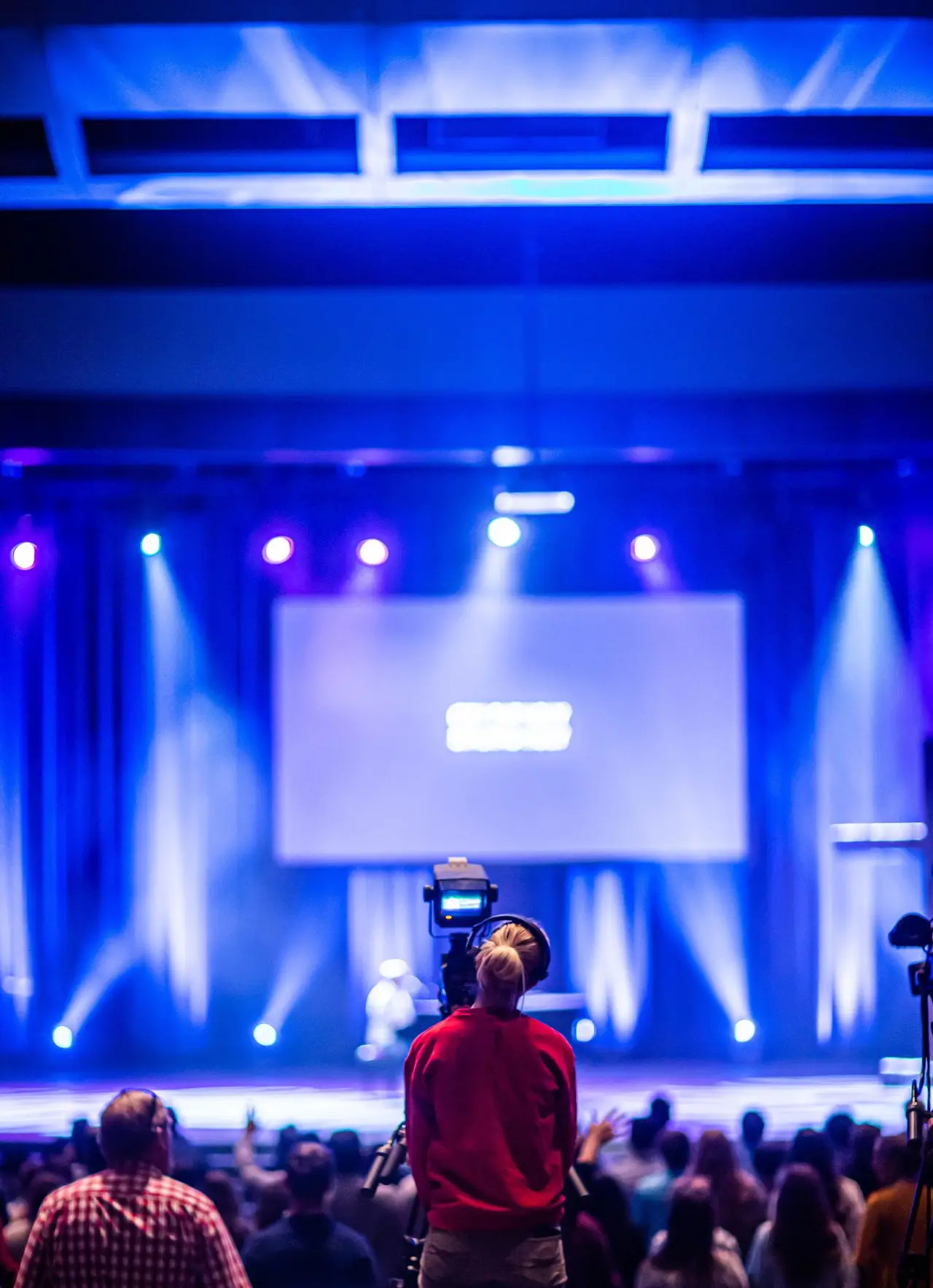 Operatore video riprende un palco con schermo e fasci blu davanti a una platea seduta