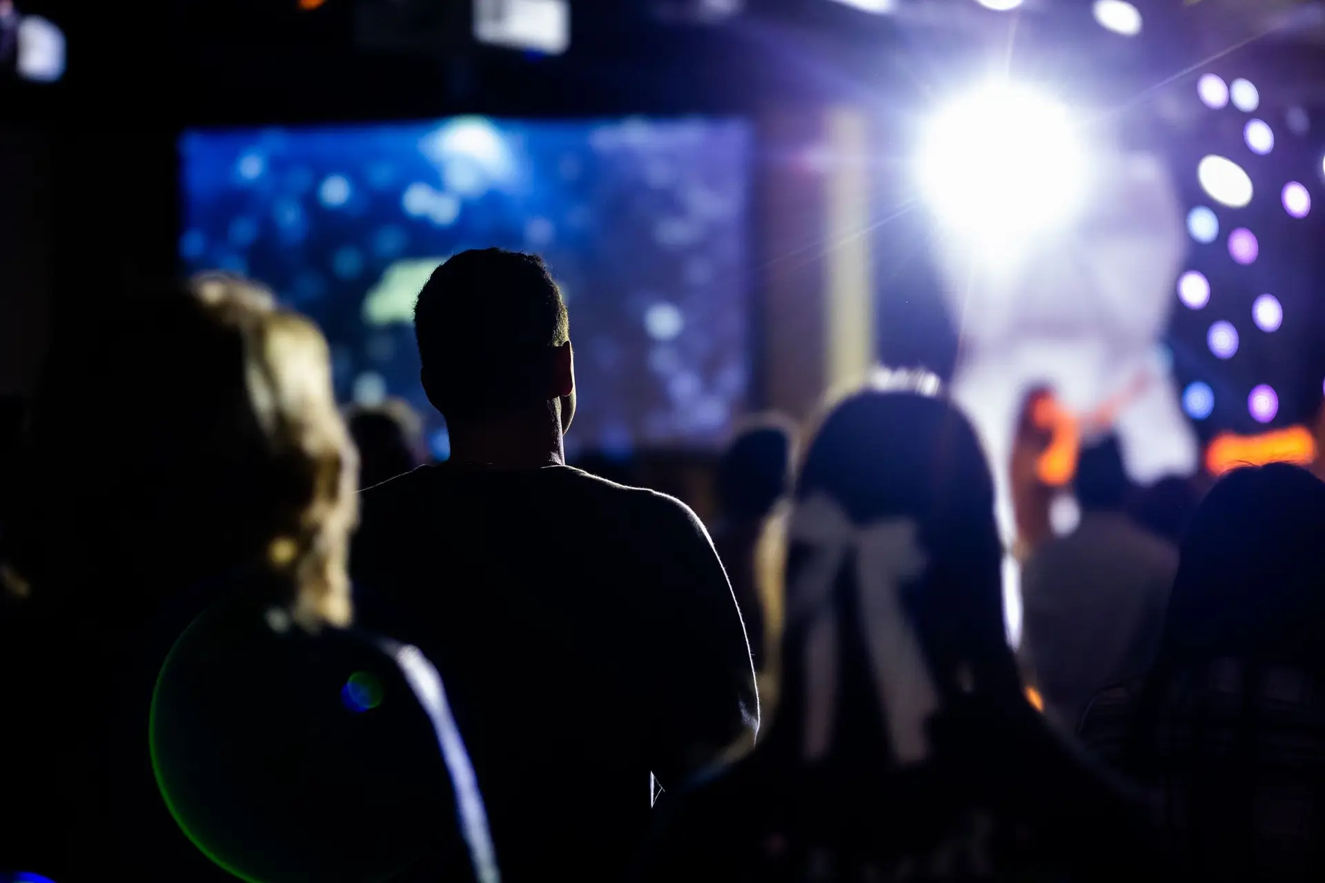 Sagome del pubblico di spalle davanti a un palco illuminato con schermo e fasci di luce